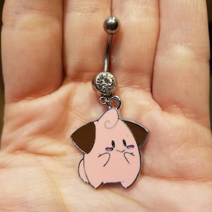 May include: A silver belly button ring with a dangling charm. The charm is a pink and brown cartoon character with a smiling expression. The ring has a clear gem and a small silver ball.
