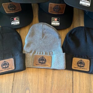 May include: A collection of black and gray beanies and black baseball caps. Each beanie and cap features a brown leather patch with a tree logo and the text "QUEEN CITY ARBORISTS". The caps have a Richardson 112 tag.