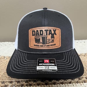 May include: Black and white trucker hat with a brown patch that reads "DAD TAX" with food graphics. The text "MAKING SURE IT'S NOT POISON" is below the main text. The hat is adjustable and has the Richardson brand label.