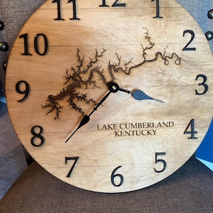May include: A round wooden clock with black numerals and hands. The clock face features a detailed laser-cut map of Lake Cumberland, Kentucky, and the text "LAKE CUMBERLAND KENTUCKY". The clock is made of light-colored wood.