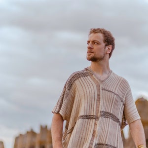 May include: A man wearing a beige and brown striped knitted poncho with a v-neck. He is standing in front of a stone wall and looking off to the side.