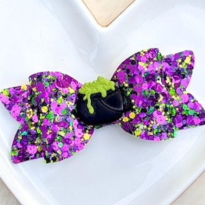 May include: A glittery purple hair bow with a black cauldron charm, filled with green bubbling potion. The bow is decorated with purple, green, and yellow glitter. A festive accessory for Halloween or themed events.