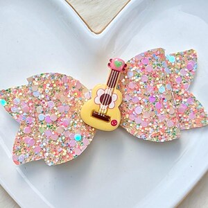 May include: A hair bow made of multi-colored glitter fabric featuring a central yellow guitar charm. The guitar charm has a white flower design on the body, a small green heart at the top of the neck, and a tiny red accent near the base of the strings.