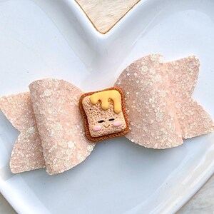 May include: A light pink glitter bow with a toast-shaped embellishment. The toast has a smiling face and is topped with yellow butter. The bow is on a white surface.