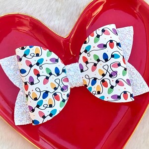 May include: A festive hair bow featuring a white background adorned with colorful Christmas lights. The bow has a glittery white accent and is displayed on a red heart-shaped dish, perfect for holiday celebrations.