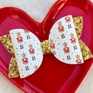 May include: A festive hair bow featuring a white background with a repeating pattern of toy soldiers in red and gold. The bow has gold glitter accents and is set on a red heart-shaped dish.