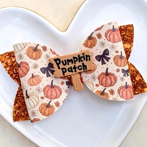 May include: A handmade hair bow with a fall theme. The bow features a cream-colored background with orange pumpkins and black bows. The bow has glittery orange accents and a small sign that reads "Pumpkin Patch".