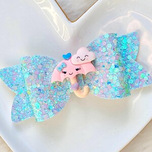 May include: A light blue glitter hair bow with a cartoon umbrella and cloud design. The bow features a pink umbrella with a smiling face, a blue bow, and a cloud with a smiling face. The bow is on a white surface.