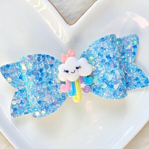 May include: A light blue glitter hair bow with a cloud and rainbow embellishment. The bow is decorated with iridescent glitter and has a small pink bow on top of the cloud. The cloud has a smiling face and colorful rainbow accents.