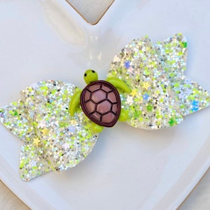 May include: A green and brown turtle charm on a white glitter bow with silver and green glitter accents. The bow is perfect for adding a touch of whimsy to any outfit.