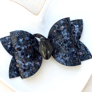 May include: A dark blue glitter bow with a black whale embellishment. The bow is made of a sparkly material and has a layered design. The whale is a glossy black color and is centered on the bow.