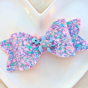 May include: A large, glittery hair bow in shades of pink, purple, and blue. A light blue turtle with pink accents is centered on the bow. The bow is on a white, heart-shaped dish.