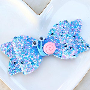 May include: A blue and purple glitter hair bow with a blue snail charm and pink shell. The snail has googly eyes.