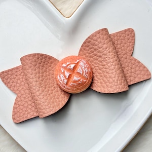 May include: A peach-colored hair bow with a textured leather-like appearance. The bow features a round, orange-colored center piece with a white, cross-shaped design. The bow is placed on a white, heart-shaped plate.