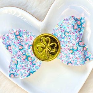 May include: A glittery hair bow with a gold coin featuring a shamrock. The bow has a light blue background with rainbow and glitter accents. The bow is on a white heart-shaped plate.