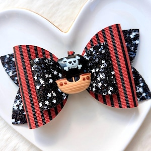 May include: A red and black striped hair bow with a black glitter and white star fabric. The bow has a small pirate ship charm with a skull and crossbones.