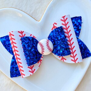 May include: A baseball-themed hair bow featuring white faux leather with red stitching, blue glitter accents, and a small baseball in the center. The bow is set against a white heart-shaped dish.