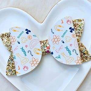 May include: A hair bow featuring a white base with a colorful science-themed print including atoms, beakers, magnets, DNA strands, and the text Science and E=MC2. The bow is accented with gold glitter fabric on the outer edges and center knot.