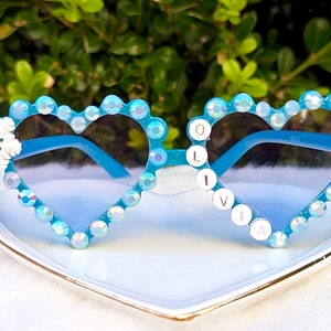 May include: Blue heart-shaped sunglasses with rhinestones and the name "Olivia" spelled out on the right lens. A white snowflake is on the left lens.