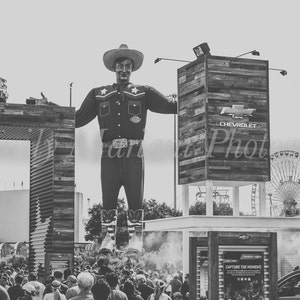 Puede incluir: Una foto en blanco y negro de un vaquero de dibujos animados grande que lleva un sombrero de vaquero y una camisa azul. Está de pie frente a un cartel de madera con el texto "Chevrolet" en él. Hay gente en el fondo.