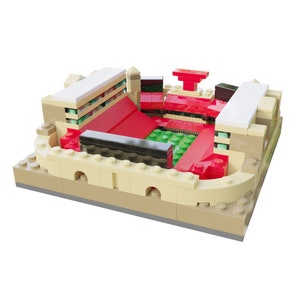 May include: A miniature stadium model constructed from interlocking plastic building bricks. The structure features tan exterior walls, red seating sections, a green field, and white roof accents. The design includes black horizontal barriers.
