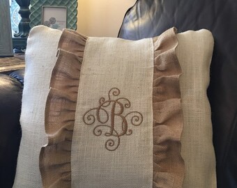 May include: Decorative square pillow with a burlap ruffle and embroidered initial "B". The pillow has a neutral color palette with a light beige background and brown accents. The pillow is on a dark brown leather surface.