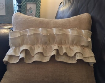 May include: A rectangular beige burlap pillow with a cream-colored ruffled trim. The pillow is placed on a dark leather couch. The pillow has a textured appearance and a decorative design.