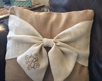 May include: A square burlap pillow with a large, cream-colored bow across the front. The bow is centered and has an embroidered monogram. The pillow is a neutral color, suitable for home decor.