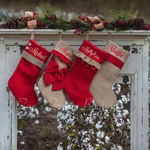 May include: Four Christmas stockings hang from a distressed white wooden frame, decorated with pine and berries. Two red stockings with names embroidered in red, and two tan stockings, one with a red bow and the other with a red ruffle.