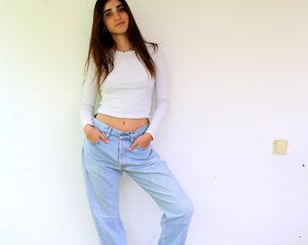 May include: A woman wearing a white long-sleeved shirt and light blue jeans with the cuffs rolled up. She is wearing brown sandals and standing in front of a white wall.