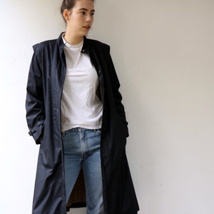 May include: A long black raincoat with a stand-up collar and a button fastening. The raincoat is worn over a white t-shirt and blue jeans.
