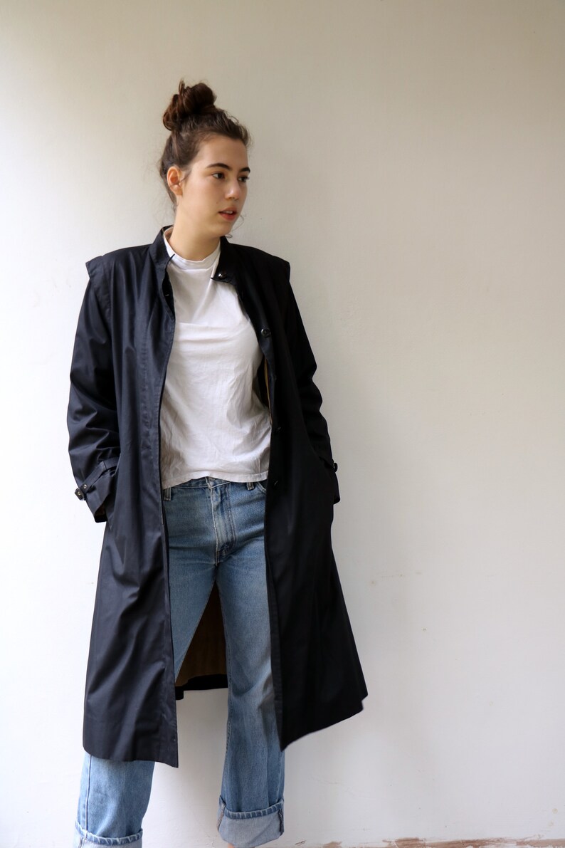 May include: A long black raincoat with a stand-up collar and a button fastening. The raincoat is worn over a white t-shirt and blue jeans.