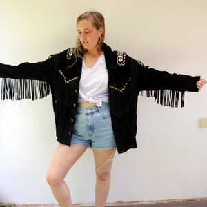 May include: A black suede jacket with fringe detailing and gold embroidery. The jacket has a button closure and is worn over a white t-shirt and denim shorts.