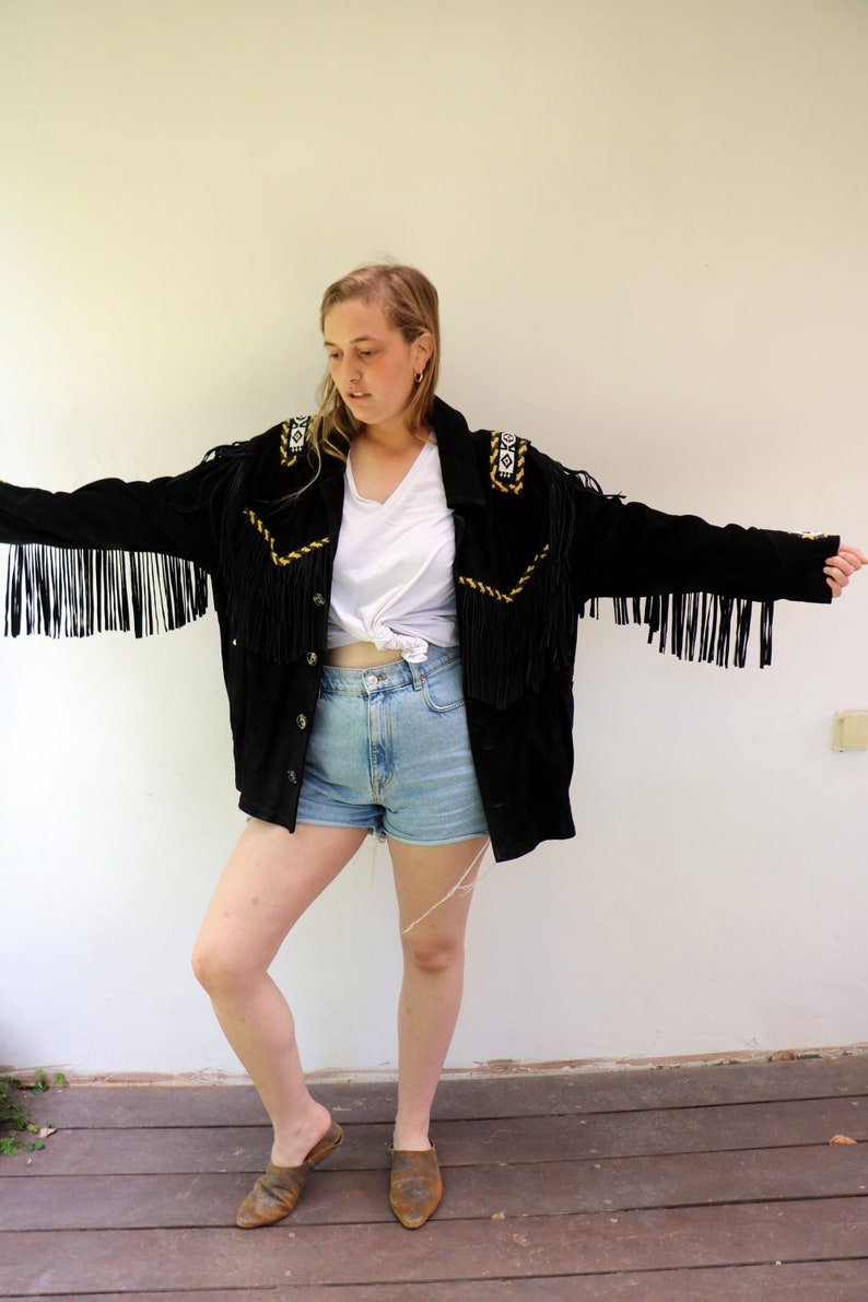 May include: A black suede jacket with fringe detailing and gold embroidery. The jacket has a button closure and is worn over a white t-shirt and denim shorts.