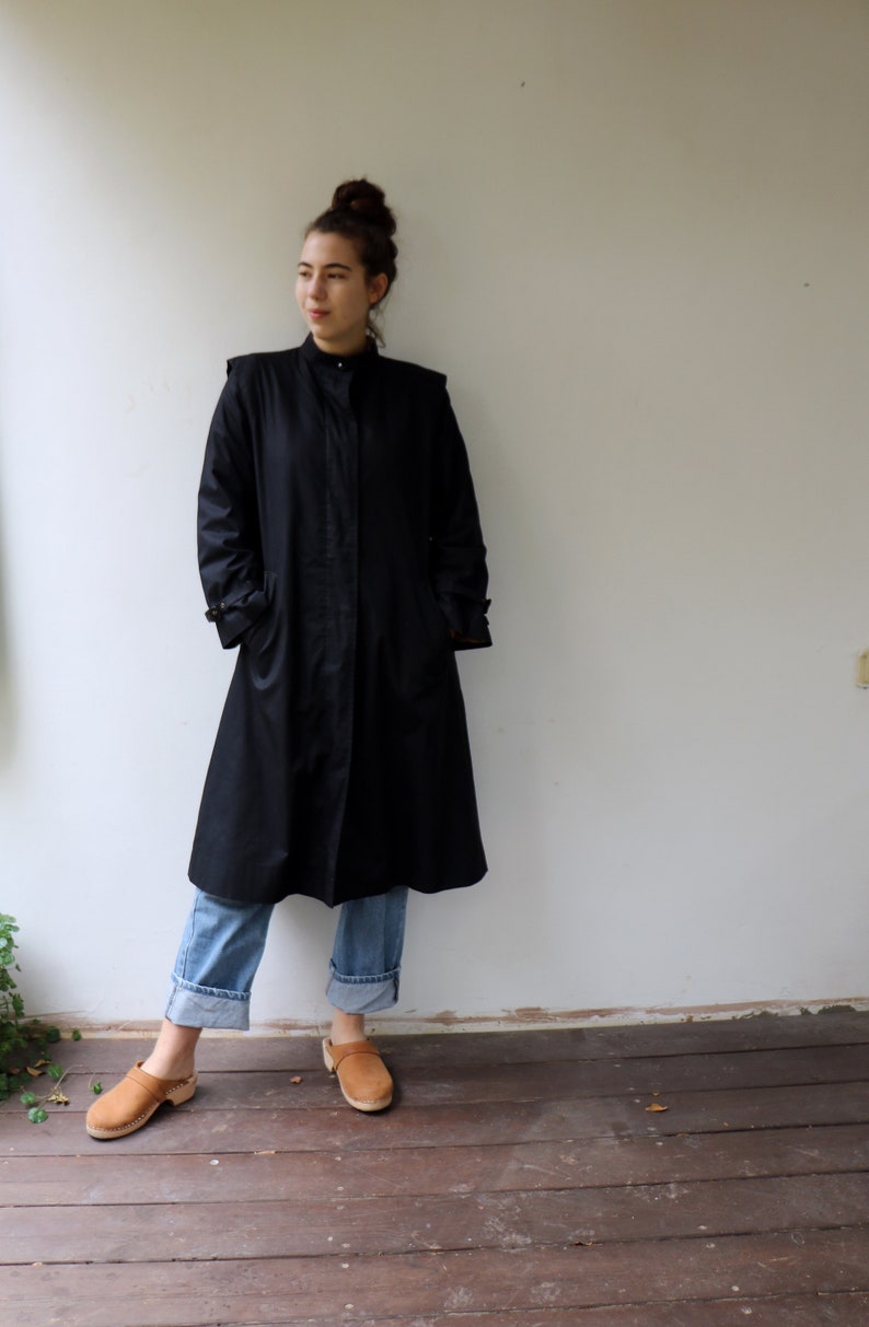 May include: A woman wearing a black long-sleeved coat, blue jeans, and brown clogs. She is standing on a wooden floor in front of a white wall.