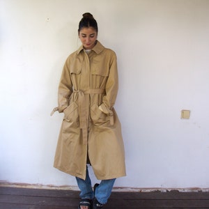 May include: A beige trench coat with a belt and pockets. The coat is being worn by a person with brown hair and is standing on a wooden floor.