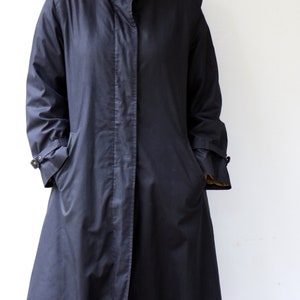 May include: A long, dark blue coat with a stand-up collar and a single button fastening. The coat has a long, flowing silhouette and features two pockets on the front.