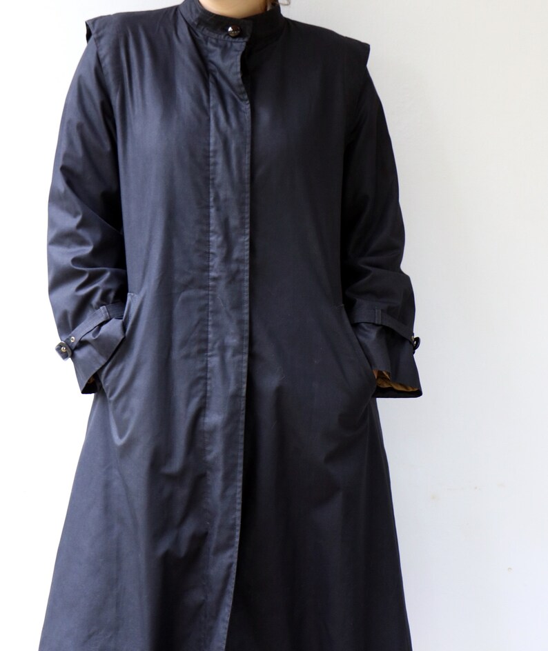 May include: A long, dark blue coat with a stand-up collar and a single button fastening. The coat has a long, flowing silhouette and features two pockets on the front.