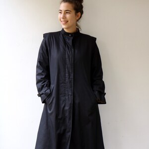 May include: A black long-sleeved coat with a high collar and a button fastening. The coat has a slightly flared silhouette and is worn over blue jeans.