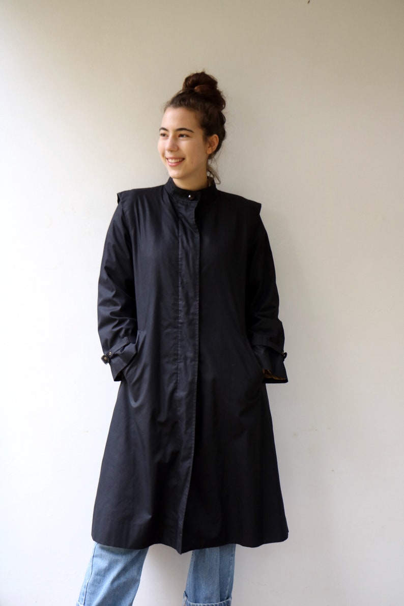 May include: A black long-sleeved coat with a high collar and a button fastening. The coat has a slightly flared silhouette and is worn over blue jeans.