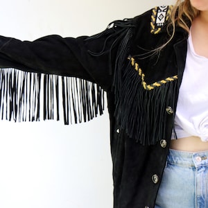 May include: A black suede jacket with fringe detailing and yellow and white embroidery. The jacket has a button closure and is worn over a white t-shirt and denim shorts.