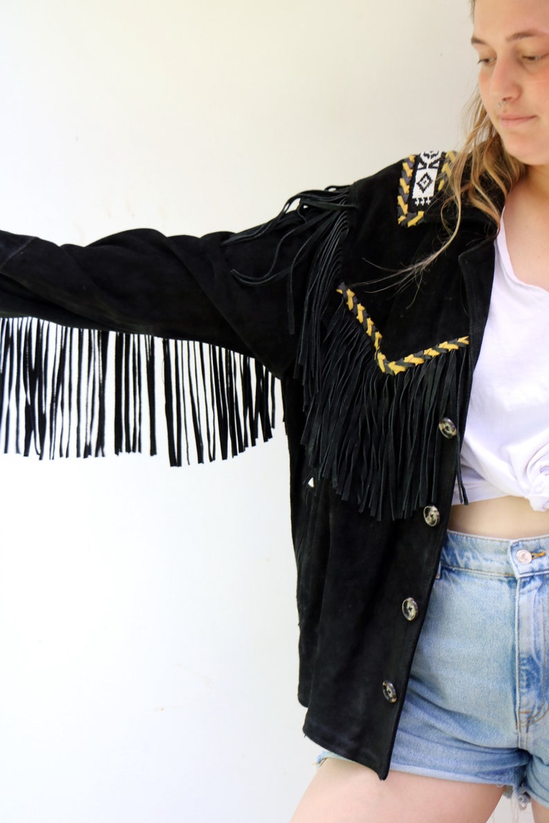 May include: A black suede jacket with fringe detailing and yellow and white embroidery. The jacket has a button closure and is worn over a white t-shirt and denim shorts.