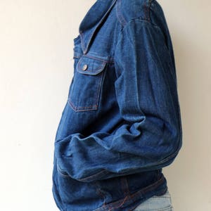 May include: A dark blue denim jacket with a collared neckline and long sleeves. The jacket has a button closure and a single chest pocket. The seams are accented with red stitching.