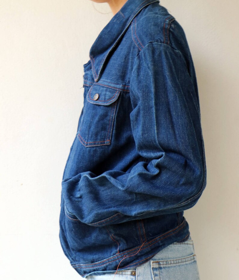 May include: A dark blue denim jacket with a collared neckline and long sleeves. The jacket has a button closure and a single chest pocket. The seams are accented with red stitching.