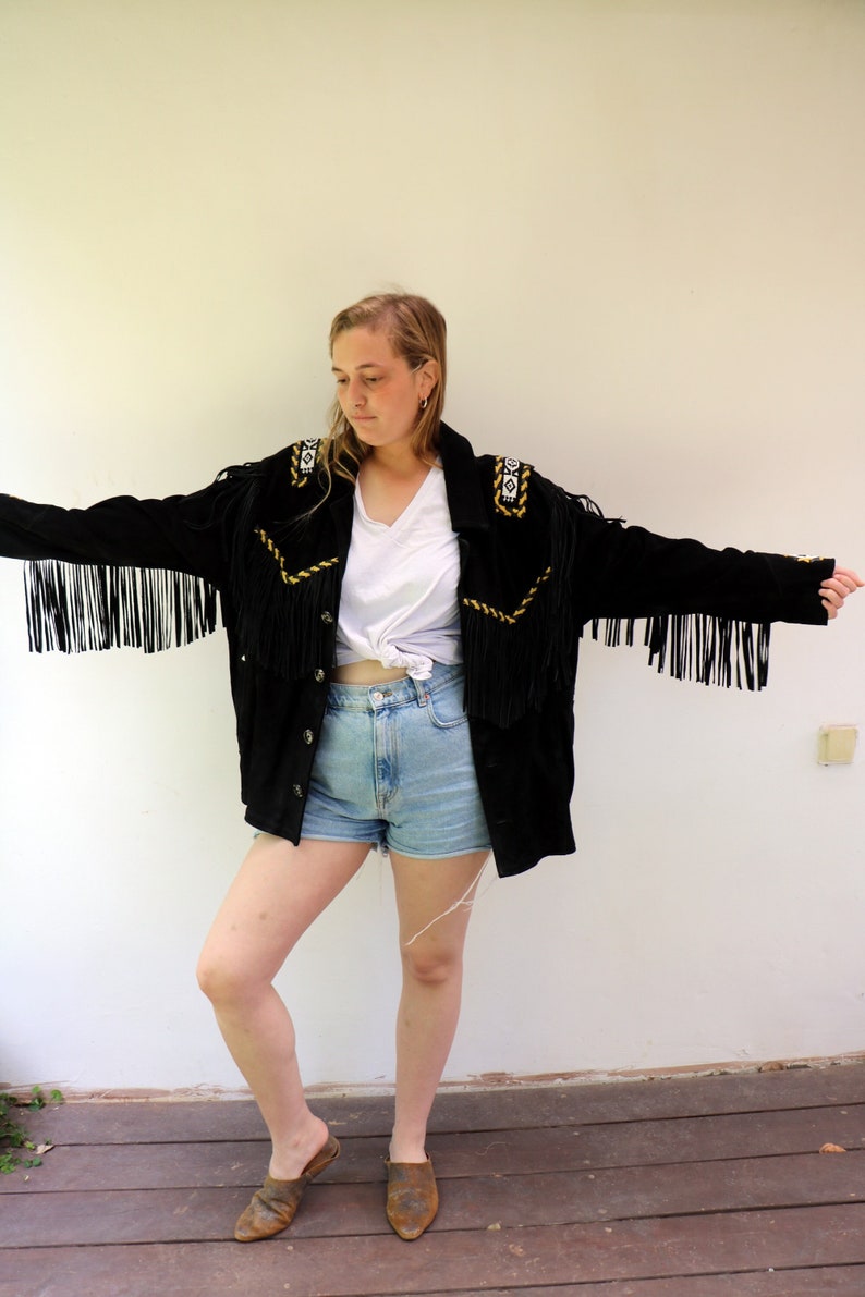 May include: A black suede jacket with fringe detailing and gold embroidery. The jacket has a button closure and a white t-shirt is worn underneath. The person is wearing denim shorts and brown leather shoes.