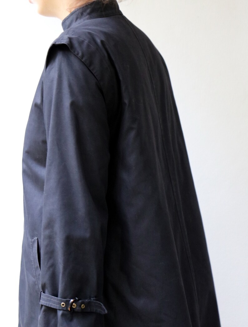 May include: A long-sleeved black jacket with a high collar and a single button fastening. The jacket has a long, flowing design and features a buckle detail on the sleeve.