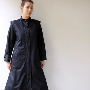 May include: A long black coat with a stand-up collar and a single button fastening. The coat has a slightly flared shape and is worn over blue jeans.