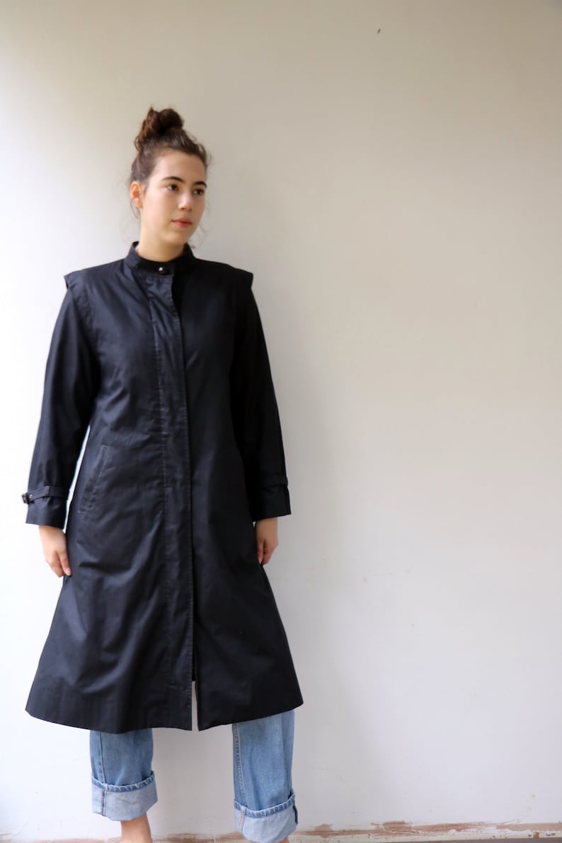 May include: A long black coat with a stand-up collar and a single button fastening. The coat has a slightly flared shape and is worn over blue jeans.