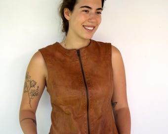 May include: A sleeveless brown leather vest with a full-length zipper. The vest is worn over blue jeans. The model has tattoos on their arms.