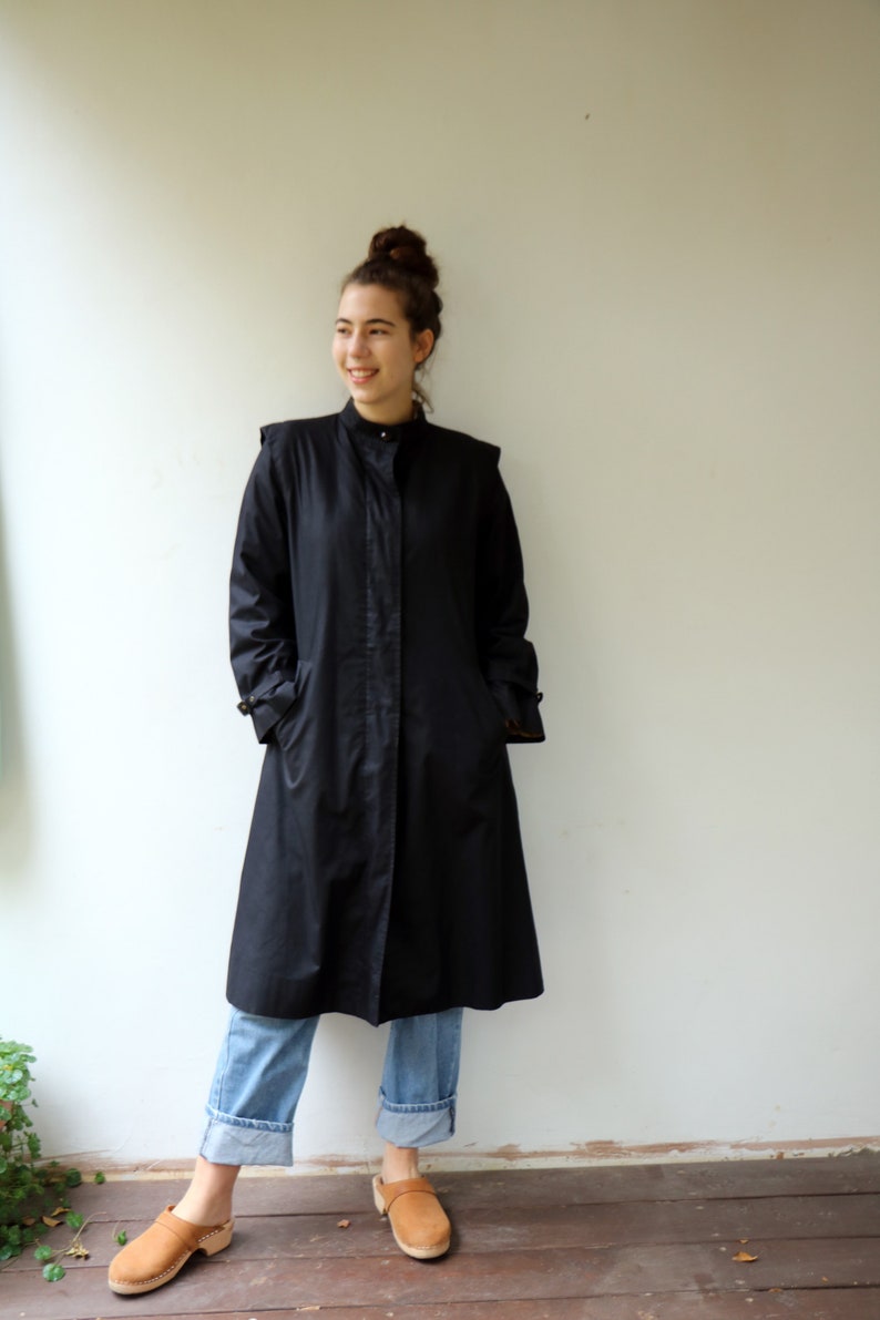May include: A woman wearing a black long-sleeved collared coat with a button closure, blue jeans, and brown clogs.